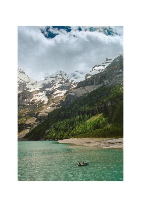 A small boat with two people floats on a turquoise lake surrounded by mountains with snow-capped peaks and a forest.