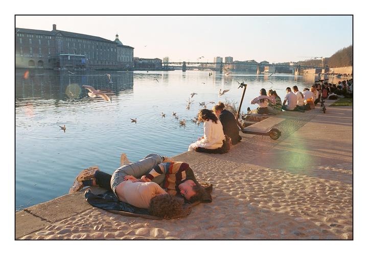 A couple lies on a stone embankment next to a river with people and birds, and a large building and bridge in the background.