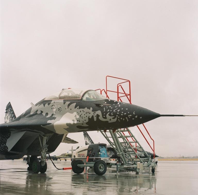 A military jet with a camouflage pattern is parked on a wet tarmac next to a red metal staircase.
