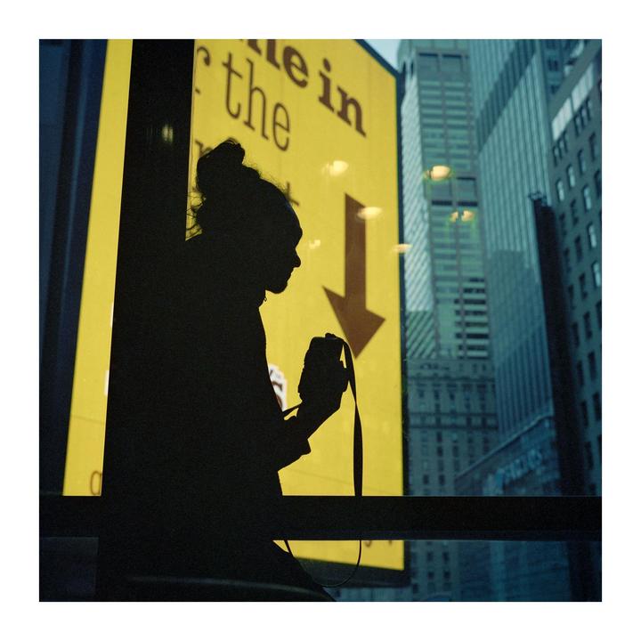 A person is silhouetted against a bright yellow sign with an arrow pointing down, with city buildings visible in the background.