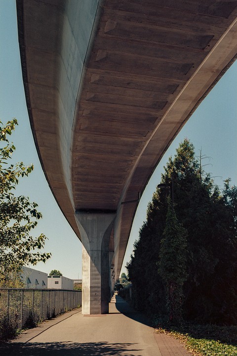 A concrete overpass curves over a paved path lined with trees and a fence.