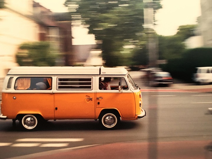 An orange and white van drives on a street with blurred buildings and trees in the background.