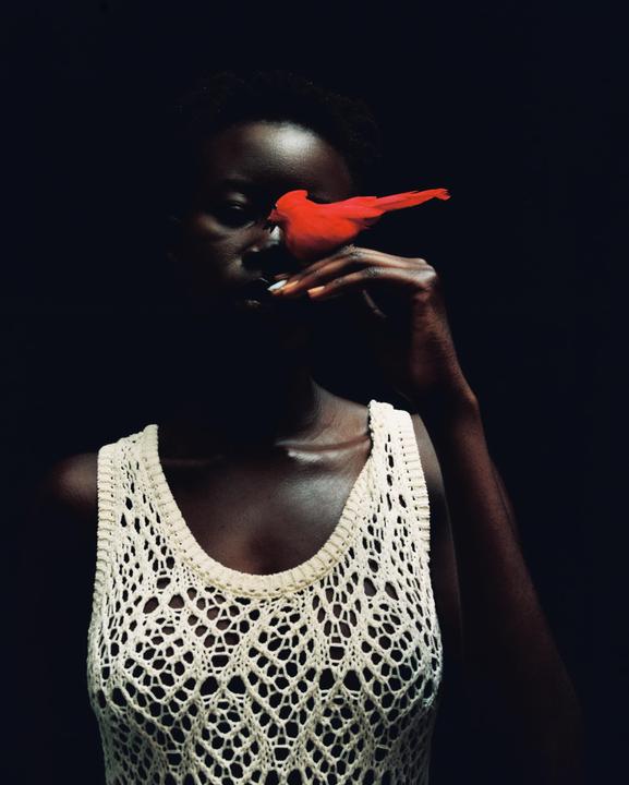 A woman in a white crocheted top holds a red bird to her face.