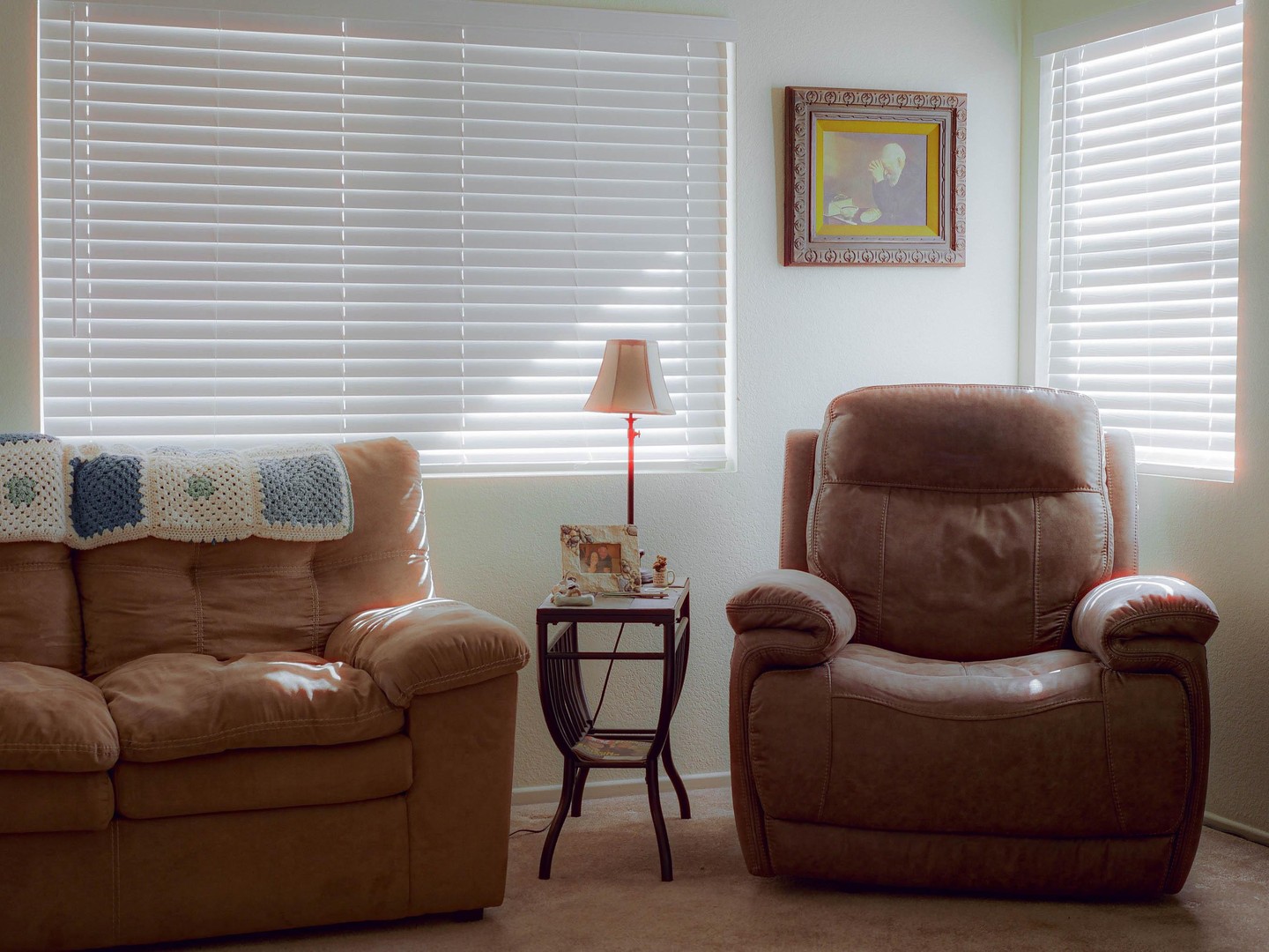 Sunlight streams through two windows with blinds, illuminating a living room with a sofa, an armchair, a side table, and a framed picture.