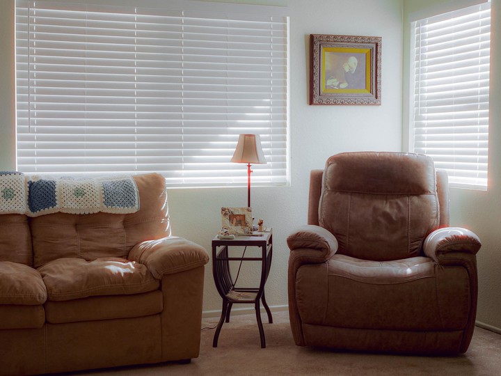 Sunlight streams through two windows with blinds, illuminating a living room with a sofa, an armchair, a side table, and a framed picture.