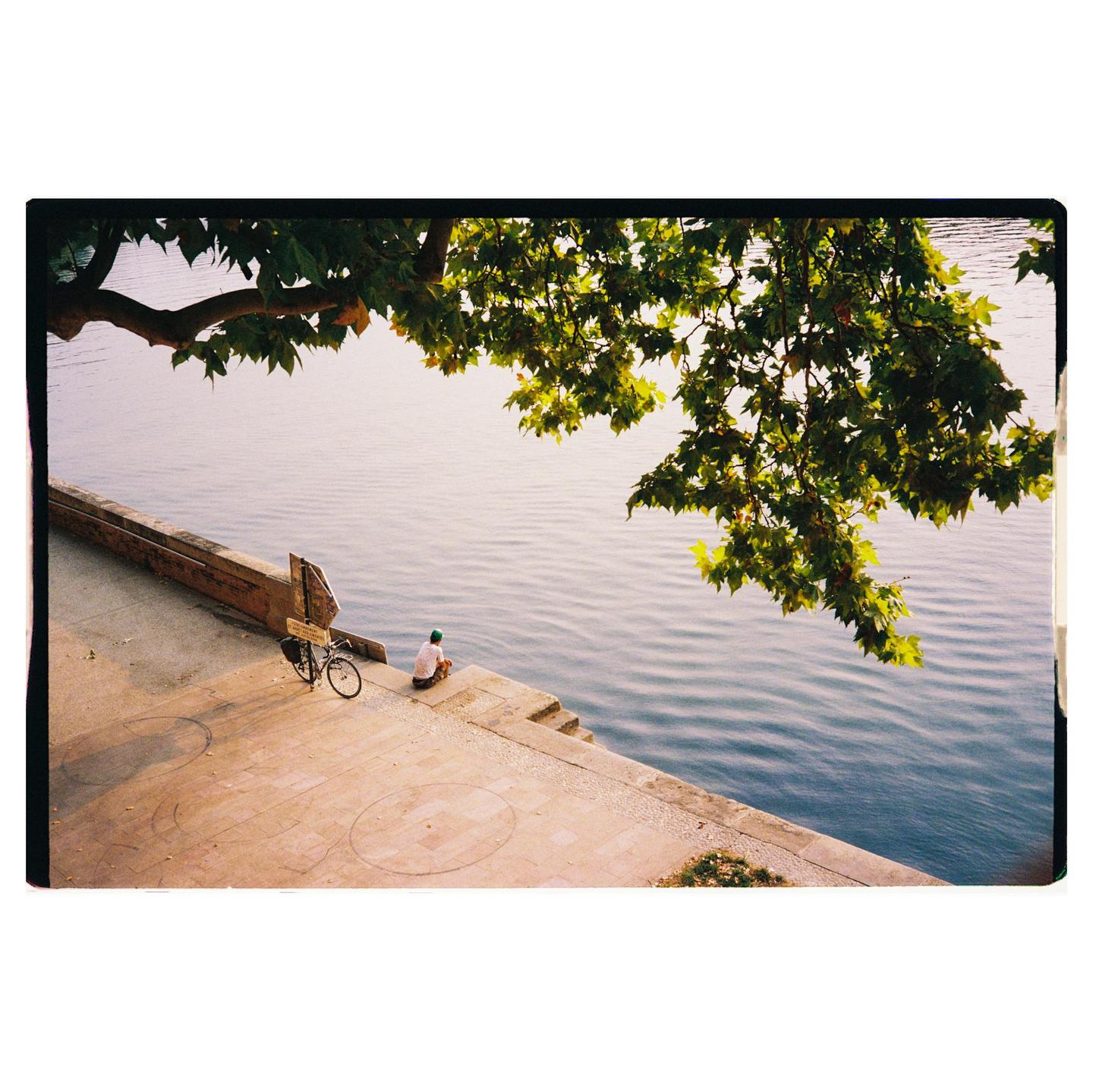 A man sits on the edge of a concrete embankment next to a body of water, with a bicycle parked nearby and a tree branch overhanging the scene.