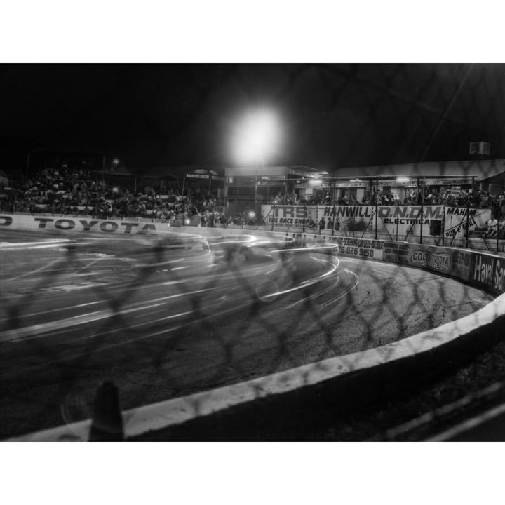 A long exposure shot captures the motion blur of race cars on a track at night, with spectators in the stands and advertisements visible in the background.