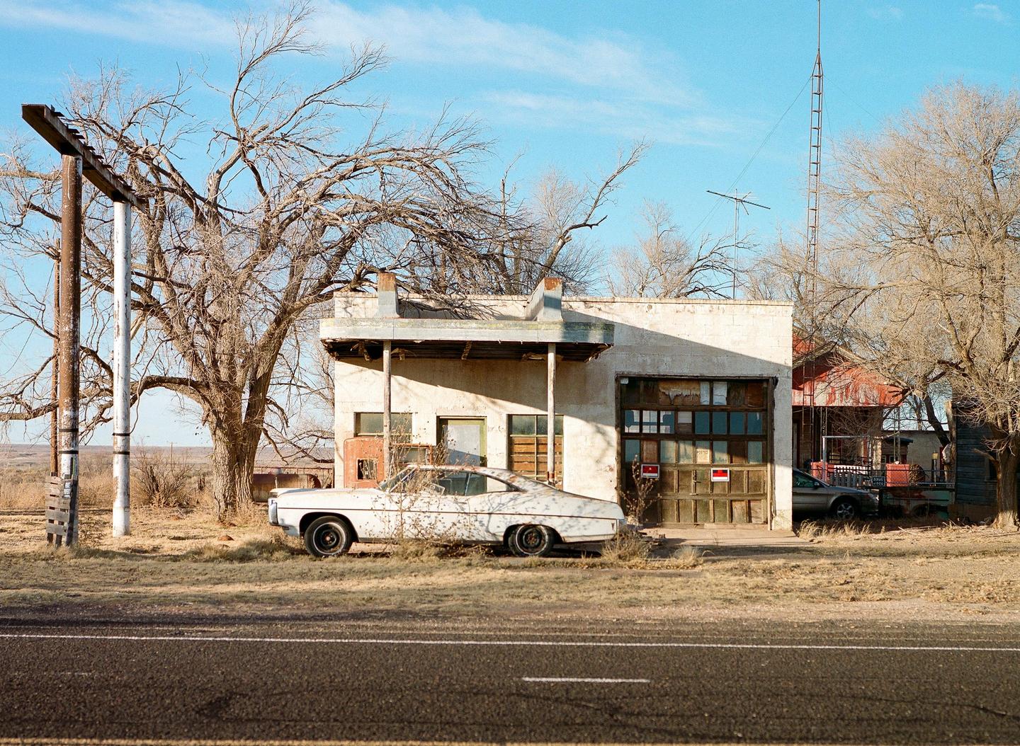 An abandoned white car is parked in front of a dilapidated building with a garage door and a covered entrance.
