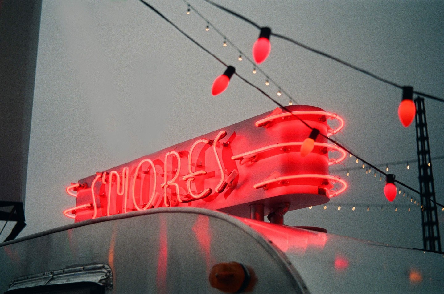 A red neon sign for "S'mores" sits atop a silver trailer, with string lights hanging above against a grey sky.