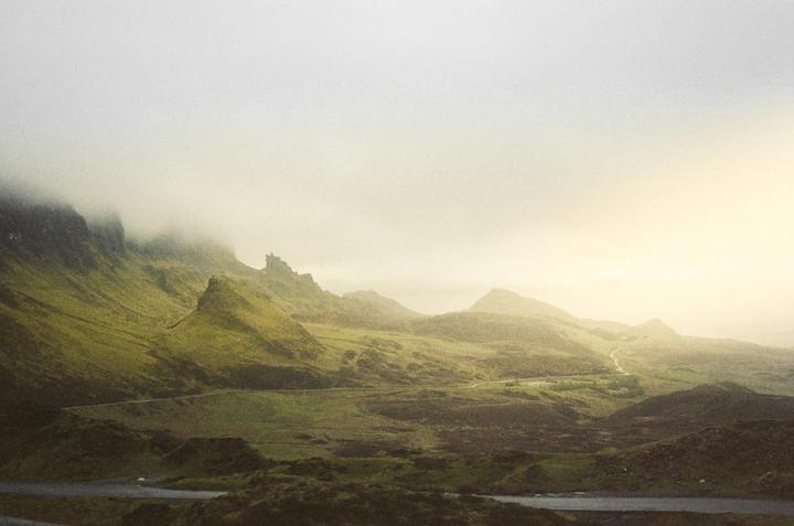 A winding road cuts through a green, mountainous landscape partially obscured by fog.