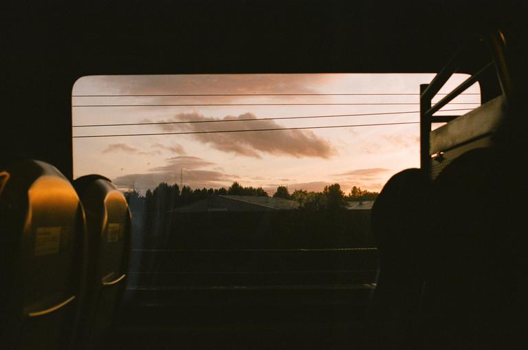 The view from a train window shows a sunset sky with clouds and power lines over a dark landscape of trees and buildings.