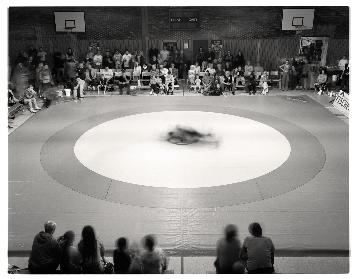 A wrestling match is taking place on a circular mat in a gymnasium with spectators watching from the stands.