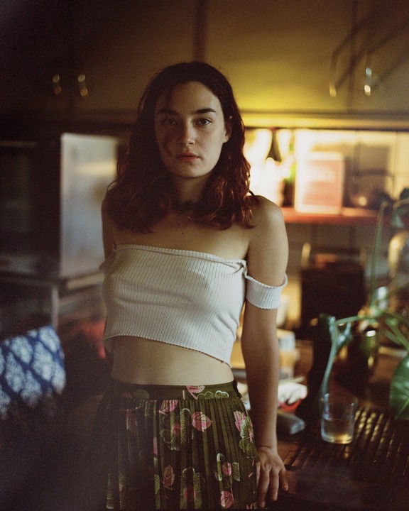 A woman with long hair and a crop top stands in a room with a patterned skirt.