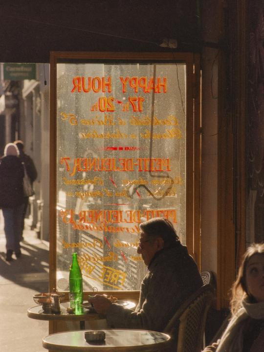 A man sits at a cafe table with a green bottle in front of a window with reversed text.