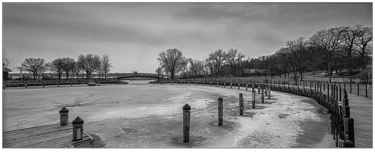 A frozen lake with a curved boardwalk and bare trees in the background is shown in black and white.