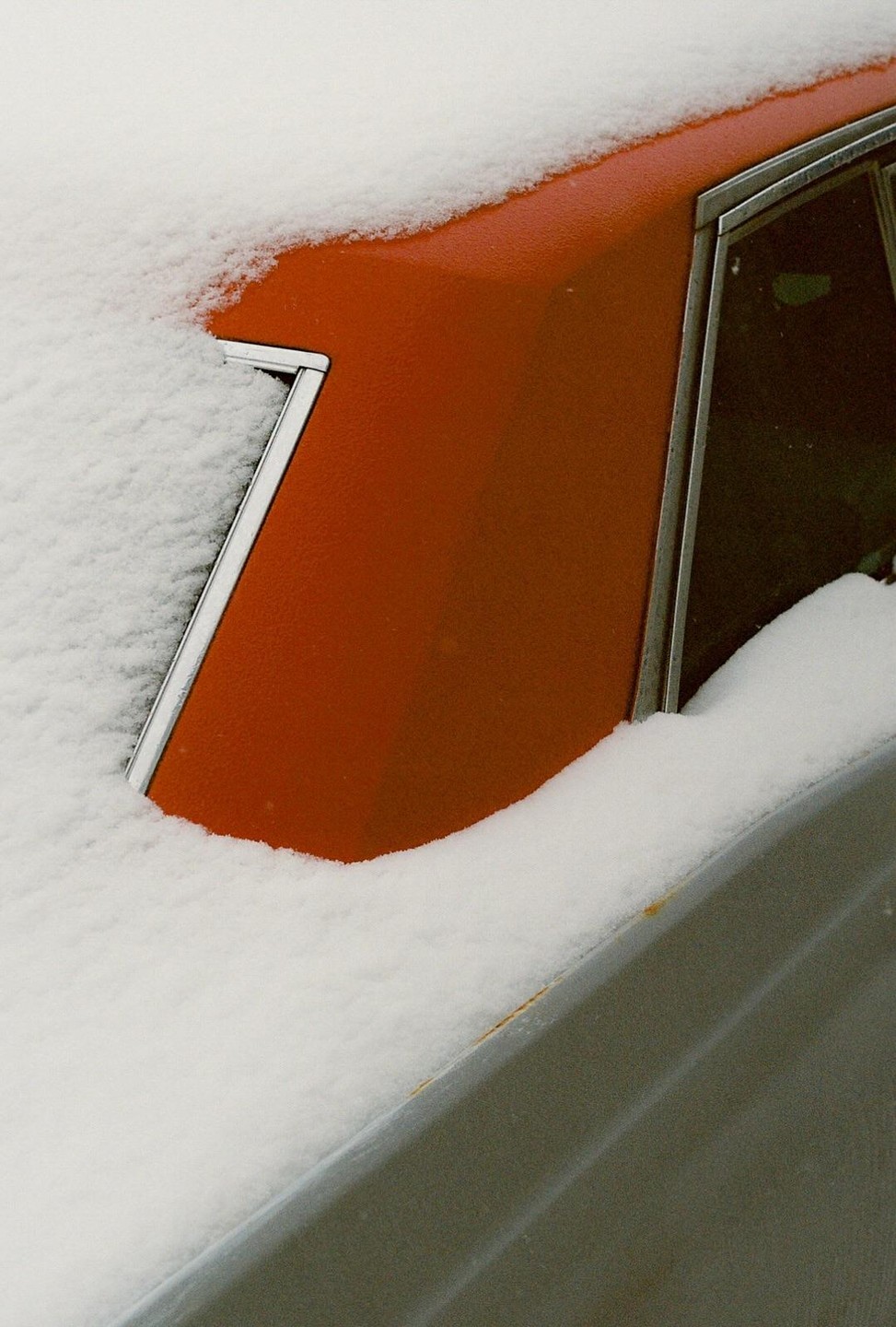 A red car is partially covered in snow, with a window visible on the right side.