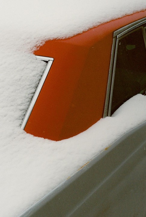 A red car is partially covered in snow, with a window visible on the right side.