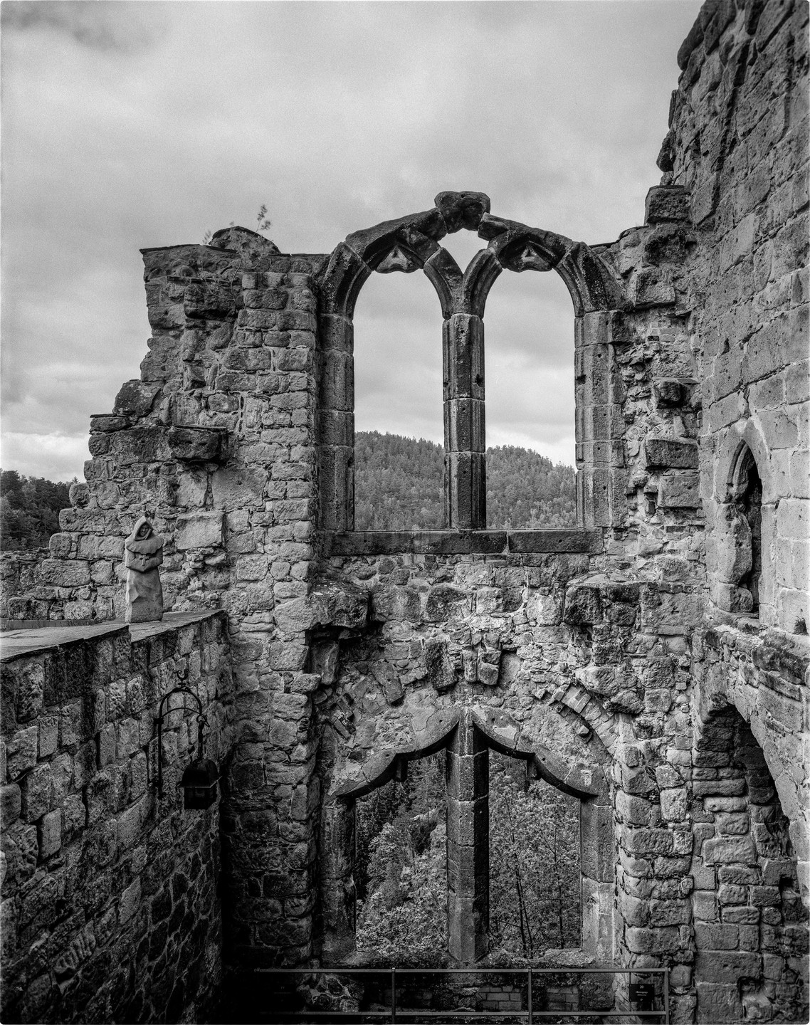 A stone statue stands on a wall overlooking the ruins of a cloister with arched windows revealing a forest and hills in the distance.