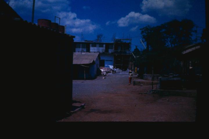 A person stands on a dirt road in a village with buildings and trees under a blue sky with clouds.
