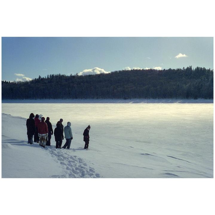 A group of people stand on a snow-covered bank next to a frozen lake with a forest in the background.