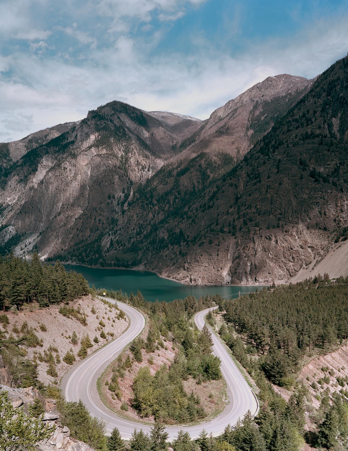 A winding road descends through a forested mountain landscape towards a lake.