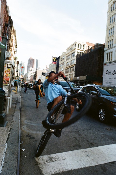 A man on a bicycle performs a wheelie on a city street with buildings and cars in the background.