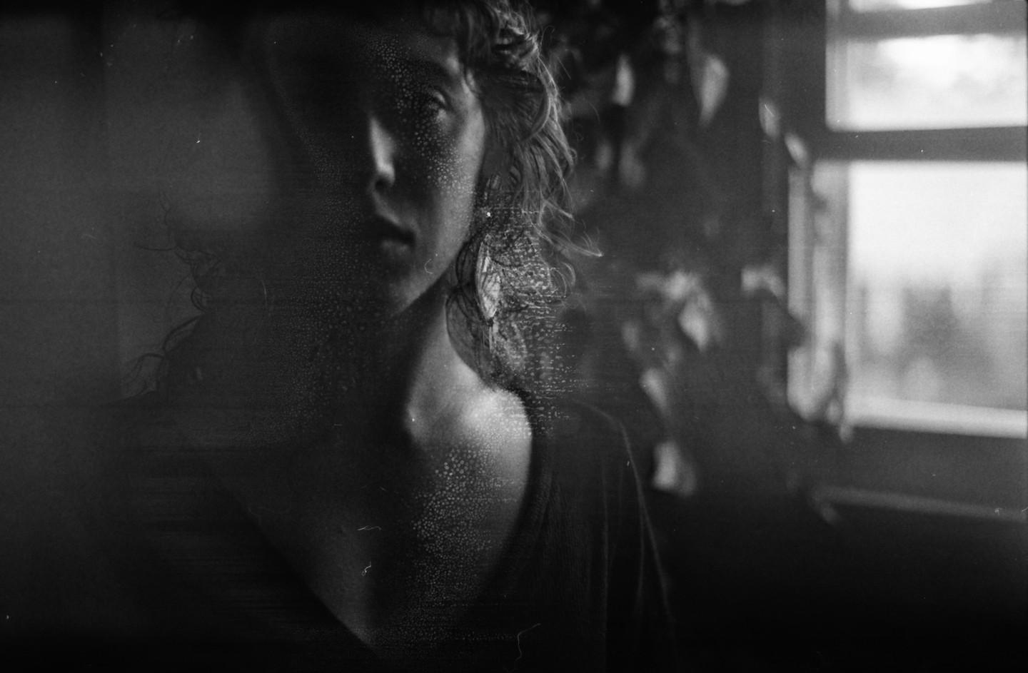 A woman with curly hair is visible in a dark room with a window and plants.