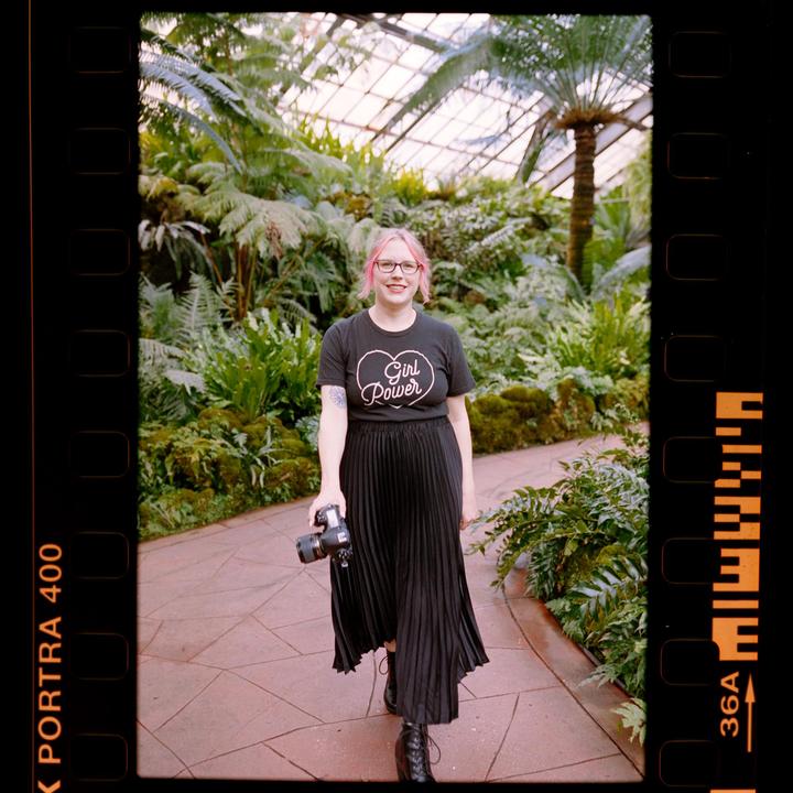 A woman with pink hair and glasses smiles at the camera while holding a camera in a lush greenhouse.