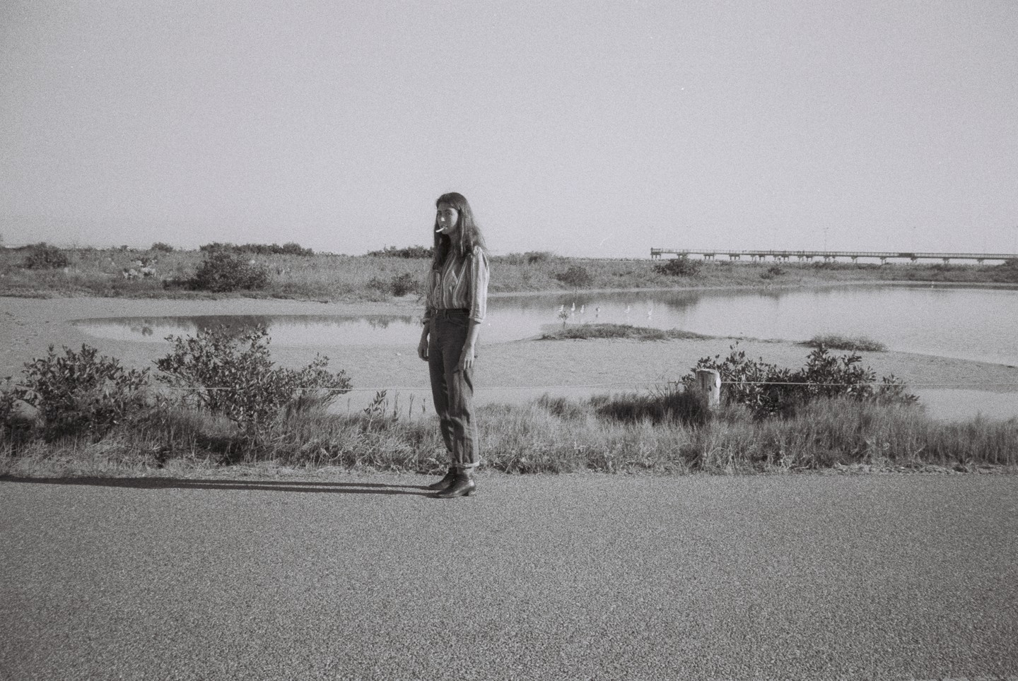 A woman stands on a paved surface next to a body of water with a long pier in the background.