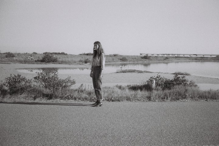 A woman stands on a paved surface next to a body of water with a long pier in the background.