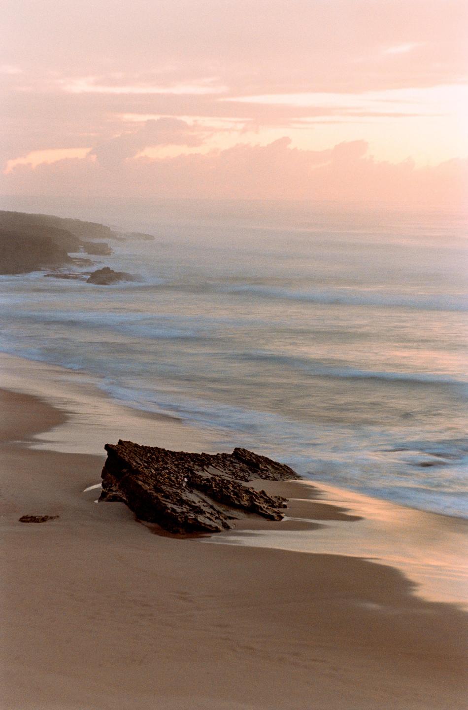 A rocky coastline with waves washing onto a sandy beach is visible under a pastel sky.