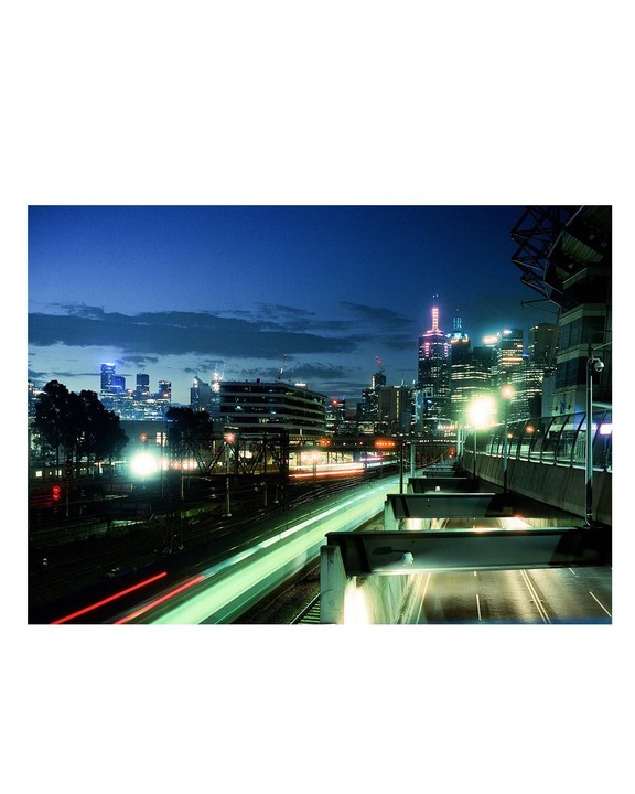 A long exposure shot captures light trails from a train and cars with a city skyline in the background at dusk.