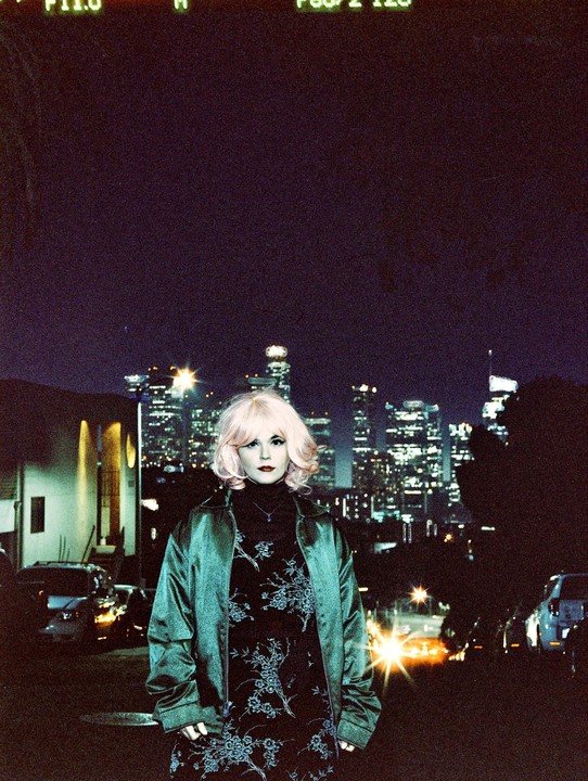 A woman with blonde hair and a green jacket stands in front of a city skyline at night.