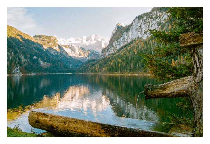 A wooden water trough with flowing water is in the foreground of a lake surrounded by mountains and forest.