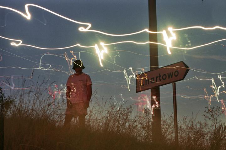 A man stands in tall grass next to a Chartowo sign with light trails streaking across the night sky.
