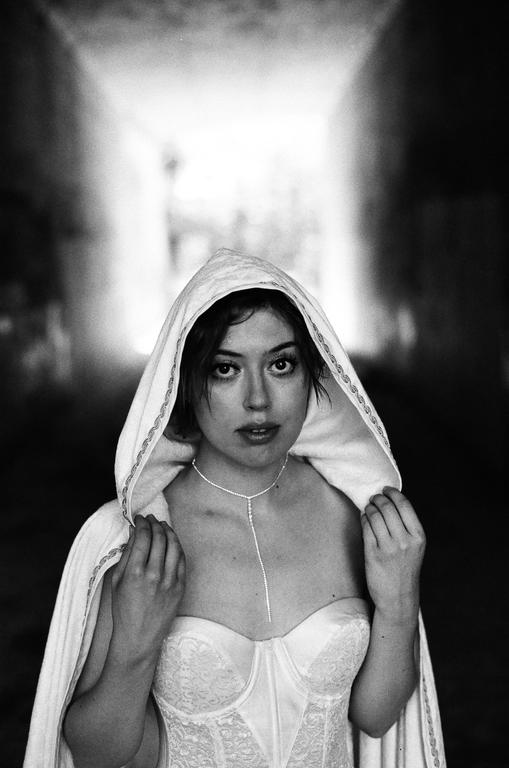A woman wearing a hooded cape and a corset stands in a tunnel.