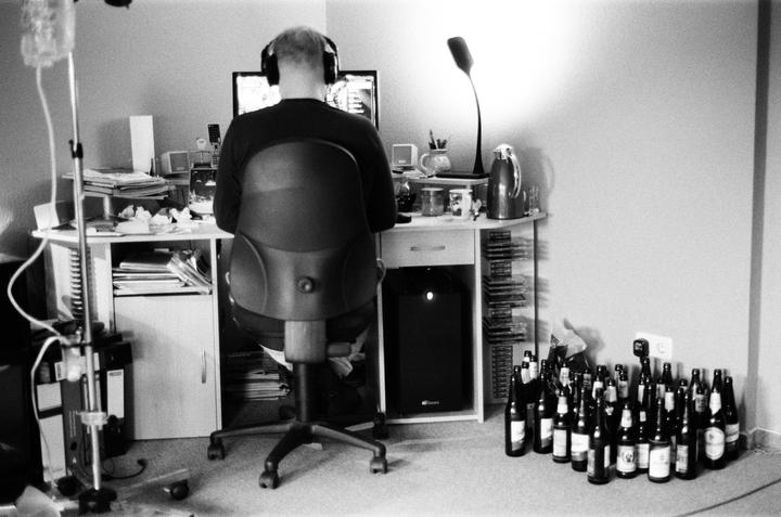A man wearing headphones sits at a cluttered desk with a computer monitor, and many empty bottles are on the floor next to the desk.
