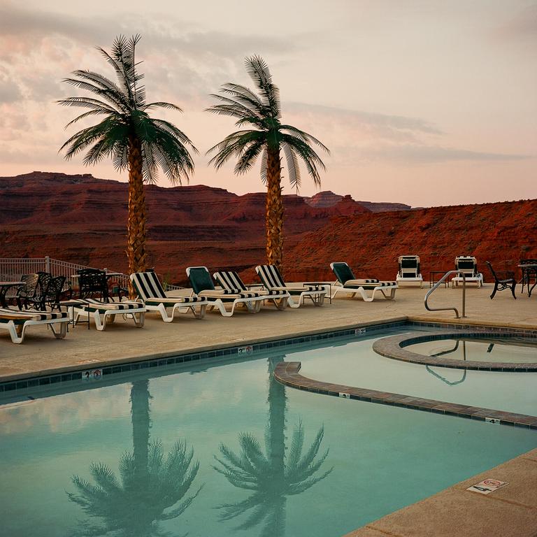 Two palm trees stand beside a swimming pool with lounge chairs, overlooking a desert landscape at dusk.