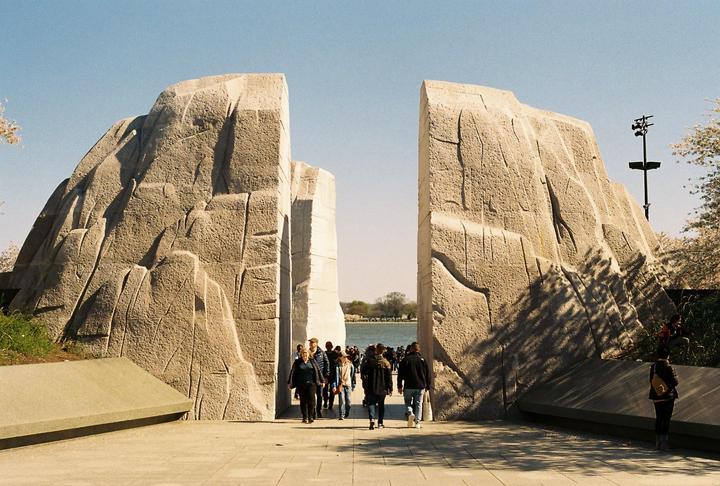 A crowd of people walk through a large stone monument with a body of water visible in the background.