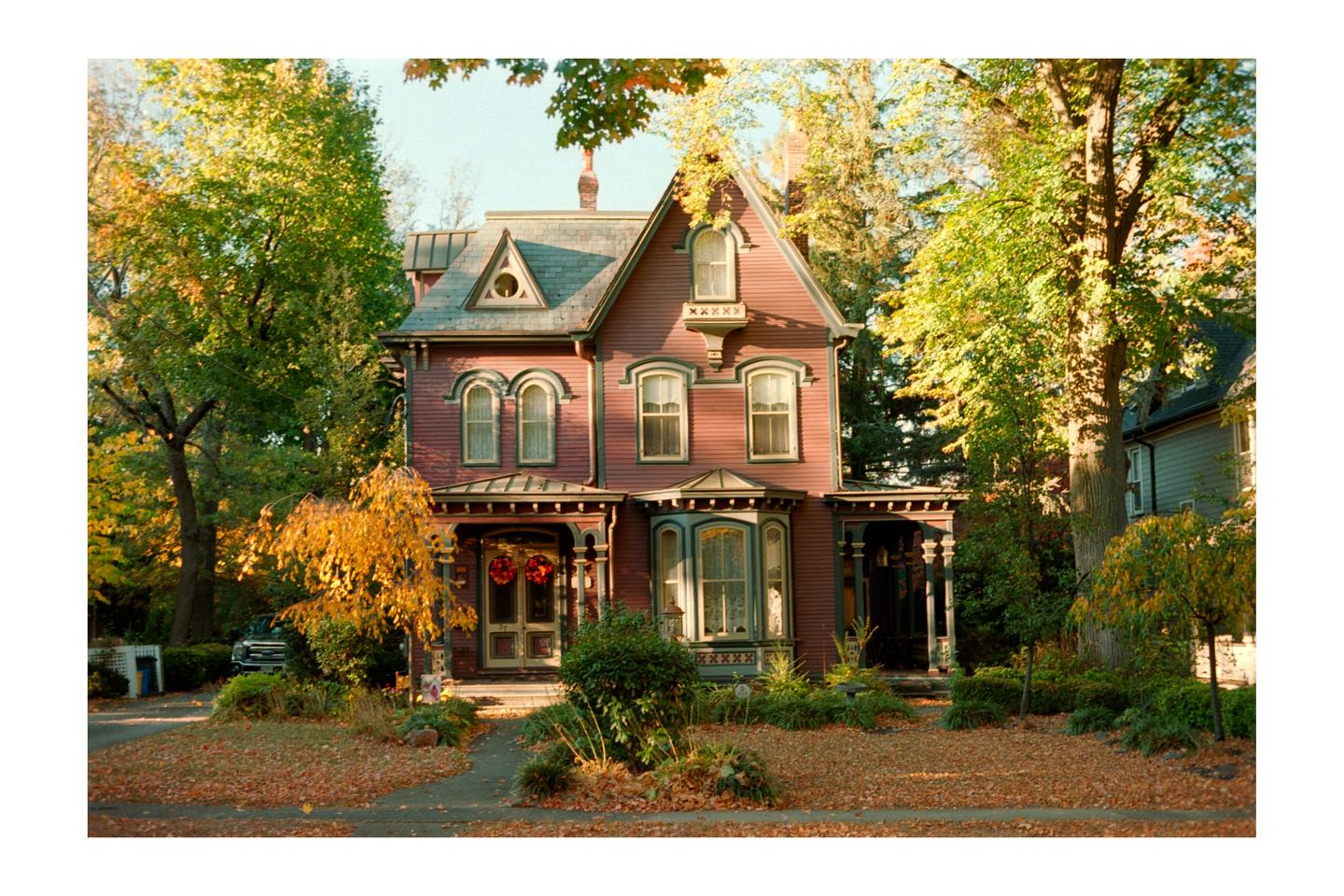 A large, ornate house with a red exterior and green trim is surrounded by trees with autumn foliage.