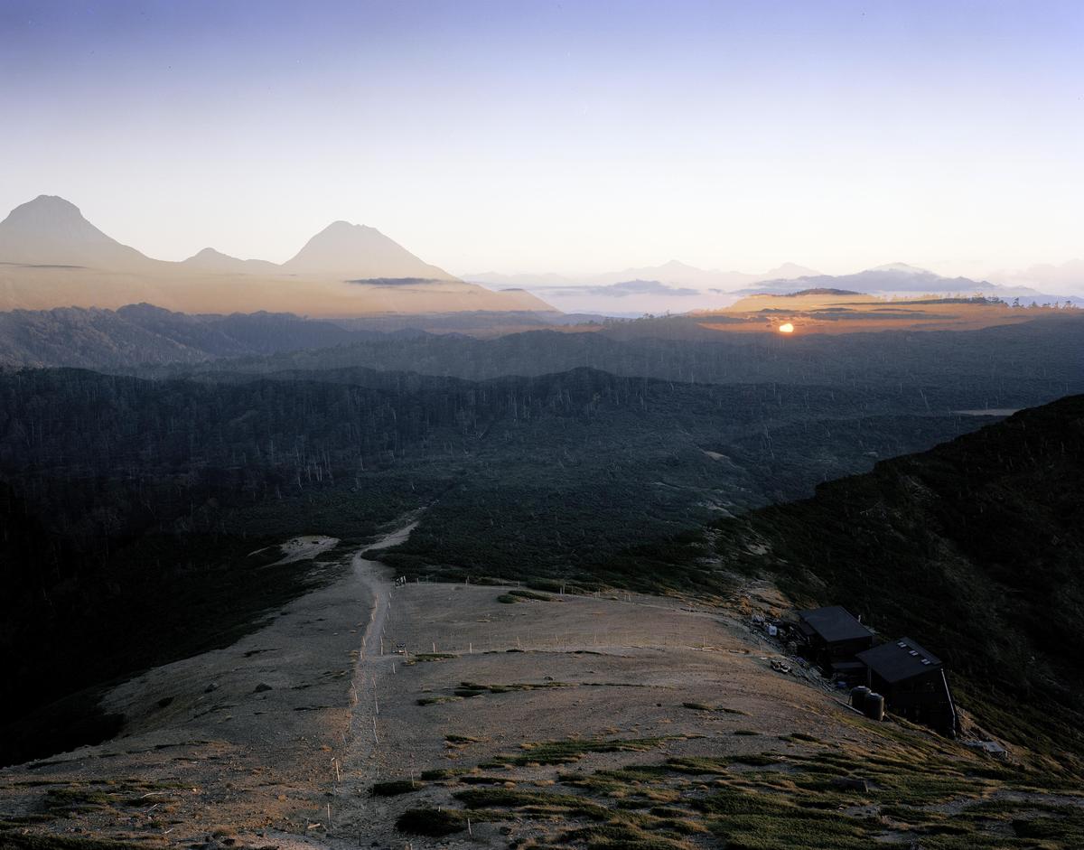 A double exposure shows a sunset over a forest and mountains, with a path leading to a building on a ridge.
