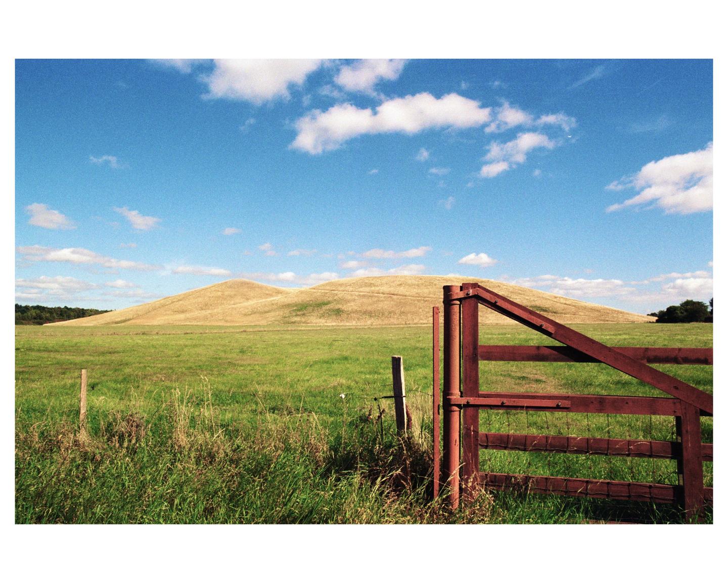 A red wooden gate stands in a green field with rolling hills under a blue sky with white clouds.