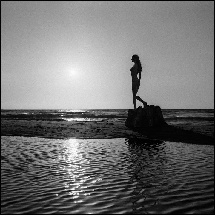 A woman stands silhouetted on a rock at the beach as the sun sets over the ocean.