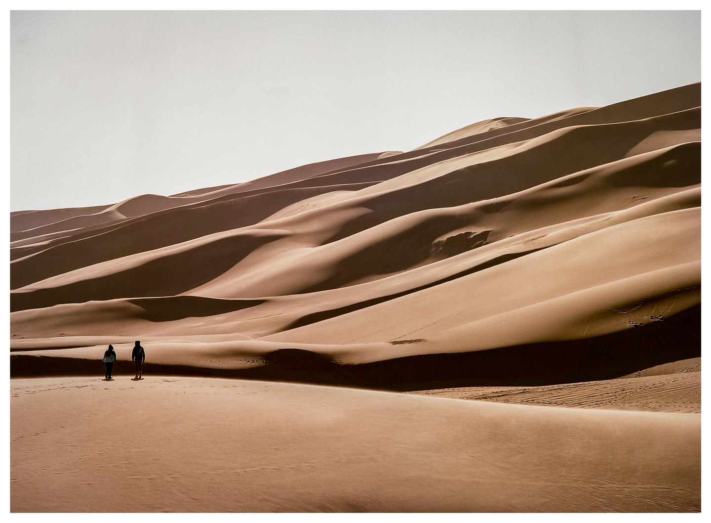 Two people stand on a vast sand dune landscape under a bright sky.