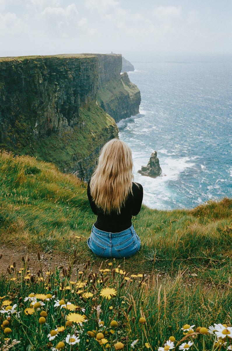 A woman with long blonde hair sits on a grassy cliff overlooking the ocean and a rocky coastline.