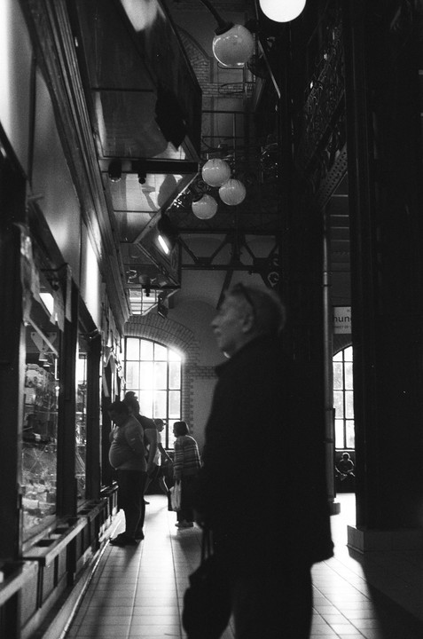 A man in profile stands in the foreground of a market hall with other people and large windows in the background.