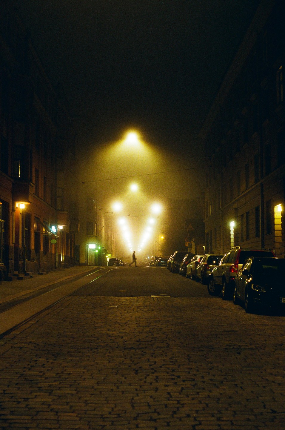 A man walks across a cobblestone street at night, illuminated by bright streetlights in the fog.