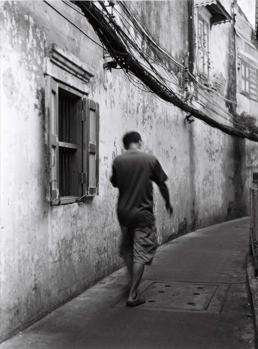 A man walks down a narrow alleyway next to a weathered building with a window and many electrical wires overhead.