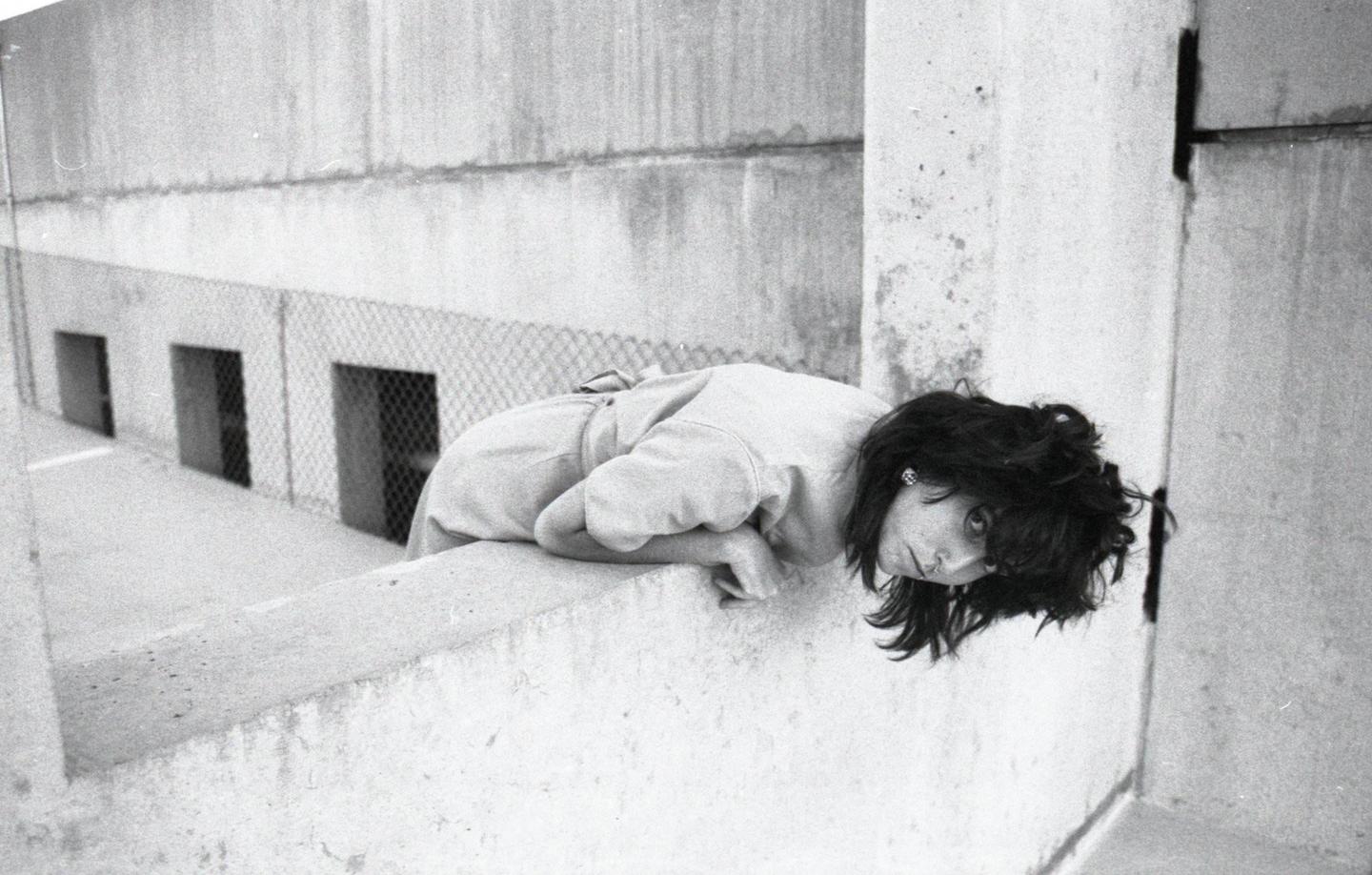 A woman leans over a concrete wall, looking up at the viewer.