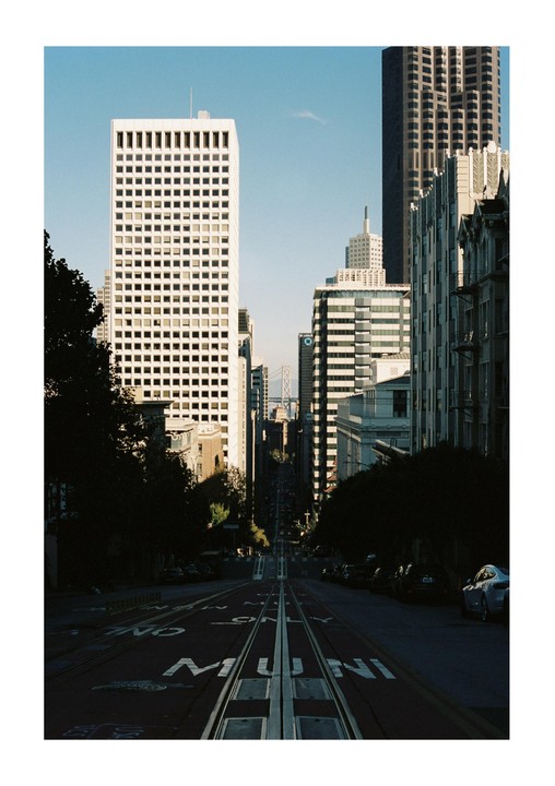 A steep street with cable car tracks leads down toward the San Francisco Bay Bridge, flanked by tall buildings and trees.